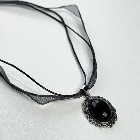 Melnā obsidiāna kulons "Ēnu vēstnesis" nofotogrāfēts slīpi uz gaišas virsmas no ginella