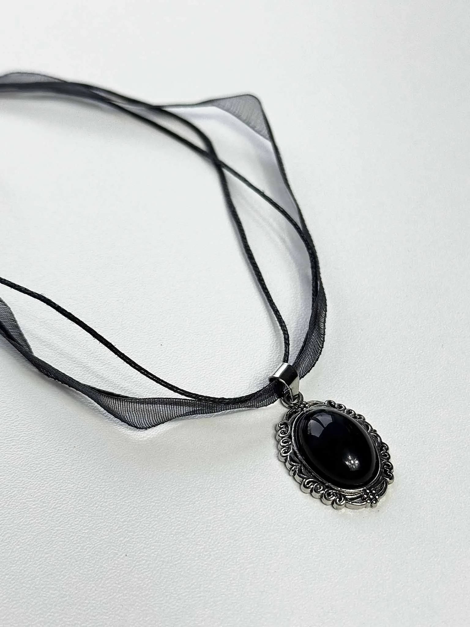 Melnā obsidiāna kulons "Ēnu vēstnesis" nofotogrāfēts slīpi uz gaišas virsmas no ginella